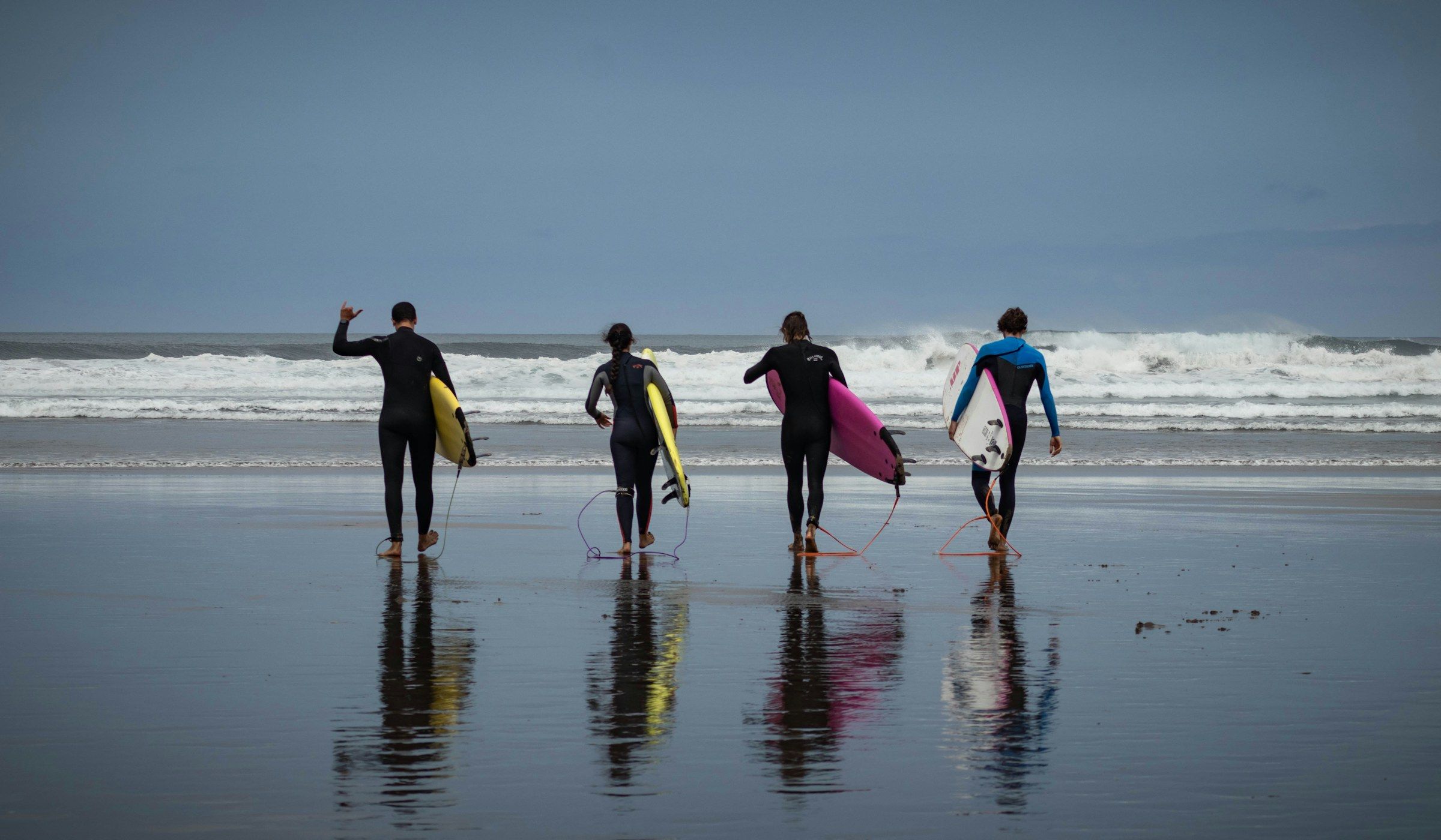 Quatre personnes en combinaison marchent vers l'océan, planche sous le bras, sur une plage mouillée
